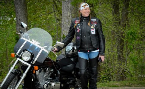 photo of a woman standing in the woods next to a motorcycle